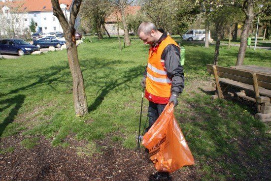 Ein Mann in einer orangefarbenen Sicherheitsweste sammelt Müll in einem Park. Er hält einen Beutel und einen Stock. Bäume, eine Bank und geparkte Autos sind im Hintergrund.