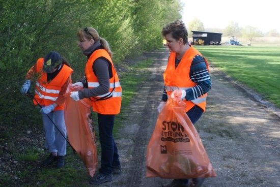 Drei Frauen in orangefarbenen Westen sammeln Müll entlang eines ländlichen Pfades, eine hält einen Stock und eine andere trägt einen vollen orangefarbenen Beutel.