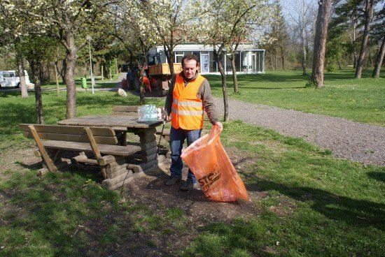 Ein Mann in einer Warnweste steht in einem Park, hält einen Beutel und hat einen Picknicktisch und eine Bank hinter sich.