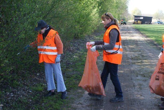 Zwei Frauen in orangefarbenen Westen sammeln Müll am Straßenrand, eine hält einen Plastikbeutel und die andere benutzt ein Werkzeug.