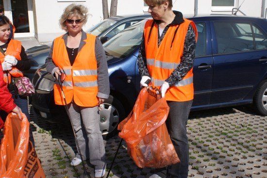 Zwei Frauen in orangen Westen und Handschuhen halten orange Taschen und stehen vor einem blauen Auto.