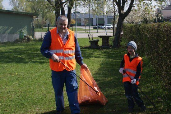 Ein Mann und ein Kind in orange Warnwesten sammeln Müll in einem Park. Der Mann hält einen Müllsack, und das Kind hält einen Stock. Hinter ihnen steht ein Busch, eine Bank und ein Tisch. In der Ferne befindet sich ein Gebäude und ein Auto.