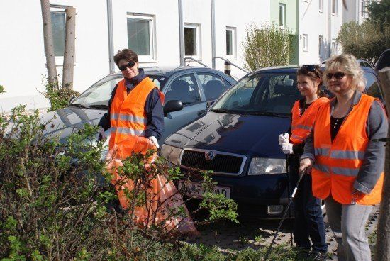 Vier Frauen in orange Weste stehen neben einem Busch und halten Müllsäcke und Stöcke. Sie sind wahrscheinlich Freiwillige, die den Bereich aufräumen. Hinter ihnen stehen zwei Autos und ein Gebäude.