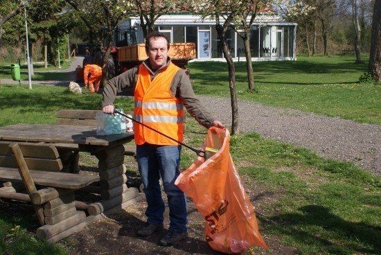 Ein Mann in einer orangefarbenen Weste hält einen Müllsack und einen Stock in der Nähe einer Picknick-Bank in einem Park.