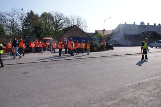 Eine Gruppe von Menschen in orangefarbenen Westen steht am Straßenrand, mit geparkten Maschinenfahrzeugen dahinter.