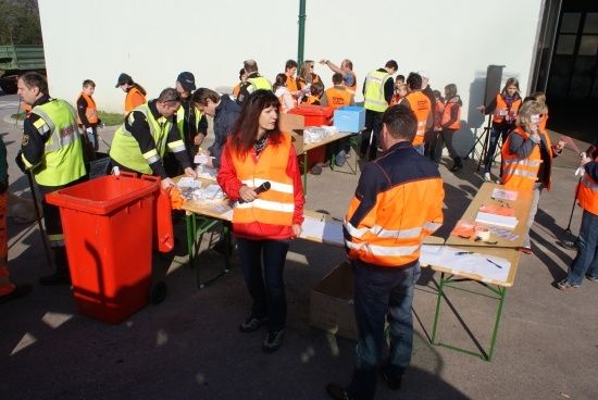 Eine Gruppe von Menschen in orangefarbenen Westen versammelt sich um Tische, möglicherweise für eine Gemeinschaftsveranstaltung. Eine Frau in einer roten Weste hält ein Mikrofon.