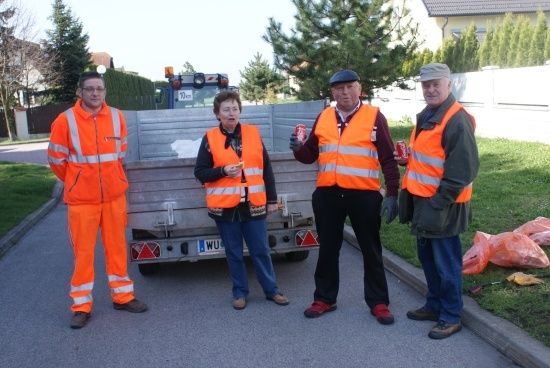 Vier Personen in orangefarbenen Warnschutzanzügen stehen am Straßenrand, zwei Männer und zwei Frauen, mit einem Lastwagen und einigen Müllsäcken im Hintergrund.