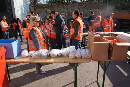 Eine Gruppe von Menschen in orangen Westen steht um einen Tisch mit verschiedenen Gegenständen in Plastiktüten. Eine Frau in der Front schaut sich die Gegenstände an.