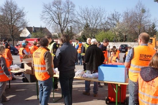 Eine Gruppe von Menschen in orangefarbenen Westen steht um Tische mit verschiedenen Gegenständen herum, möglicherweise an einer Gemeinschaftsveranstaltung teilnehmend. Bäume und Gebäude sind im Hintergrund zu sehen.