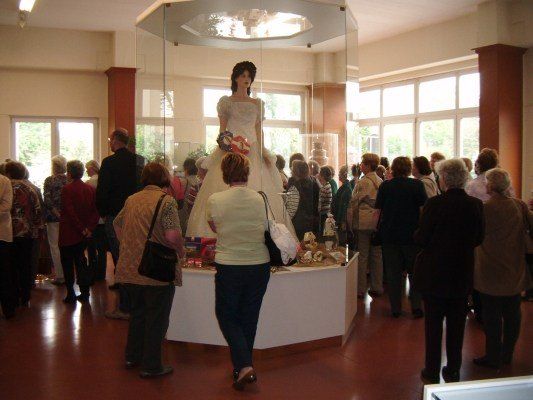 Eine Gruppe von Menschen versammelt sich um eine Glasvitrine mit einer Hochzeitskleid-Mannequin, wobei natürliches Licht durch große Fenster einströmt.