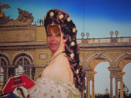 Eine Frau in historischer Kleidung mit einem Blumenkopfschmuck steht vor einem klassischen Gebäude. Sie hält eine Schachtel Sissi Taler.