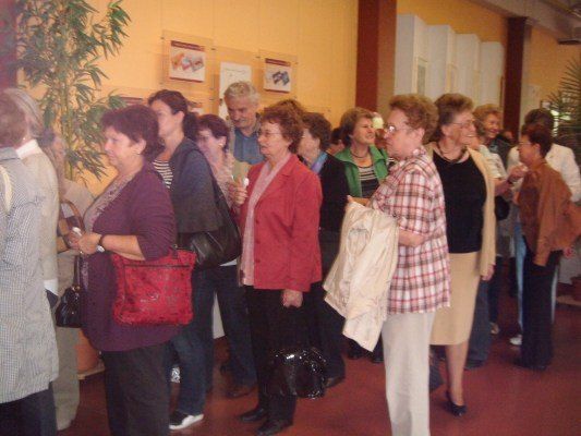 Eine Gruppe älterer Frauen und Männer steht in einer Reihe, wahrscheinlich in einer Warteschlange, in einem Raum mit gelben Wänden und Topfpflanzen.