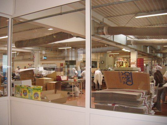 Ein Fabrikinnenraum mit Arbeitern in weißen Uniformen und Haarnetzen, die Kisten und Produkte handhaben. Der Raum ist gut beleuchtet mit Leuchtstofflampen und hat einen großen Stapel von Kisten, die mit 'CHNO' beschriftet sind.