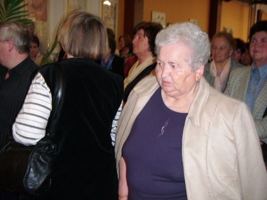 Eine ältere Frau mit weißen Haaren steht in einem Raum mit anderen Menschen, möglicherweise bei einer gesellschaftlichen Veranstaltung. Sie trägt ein lila Shirt und eine beige Jacke.