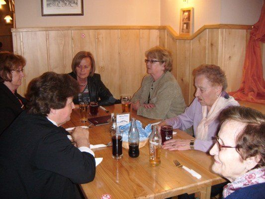 Eine Gruppe von Frauen sitzt um einen Holztisch herum, wahrscheinlich in einem Restaurant. Sie führen ein Gespräch. Der Tisch ist mit Gläsern, Flaschen und Servietten gedeckt. Die Wand hinter ihnen ist aus Holz mit Bilderrahmen.