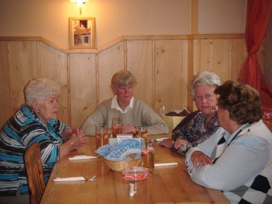 Fünf ältere Frauen sitzen um einen Holztisch herum und unterhalten sich. Gläser mit Bier und ein Korb mit Servietten stehen auf dem Tisch. Der Raum hat Holzwände und einen Bilderrahmen an der Wand.