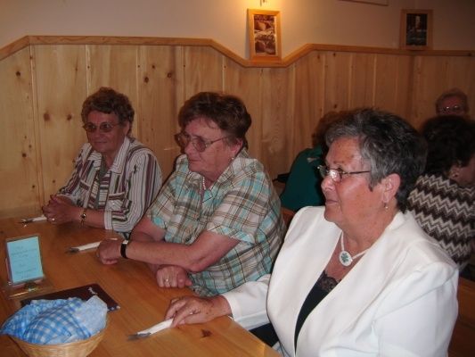 Drei Frauen mit Brille sitzen an einem Tisch in einem holzgetäfelten Raum, eine trägt einen weißen Blazer und eine andere ein kariertes Hemd.