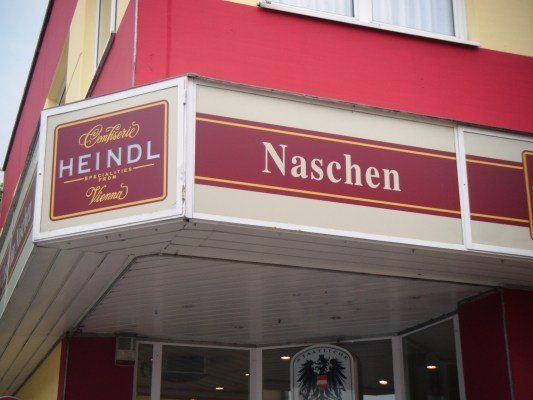 Ein Ladengeschäft mit einem roten und weißen Schild, das 'Naschen' und darunter ein kleineres Schild, das 'Heindl Confiserie Spezialitäten aus Wien' anzeigt. Der Laden hat eine Glastür und ein Fenster und ein Logo mit einem Adler auf der rechten Seite.