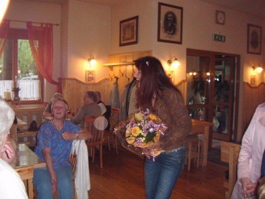 Eine Frau mit einem Blumenstrauß geht durch ein Restaurant mit Holzböden, wo mehrere Personen an Tischen sitzen. Die Wände sind mit Bilderrahmen und einer Uhr geschmückt.