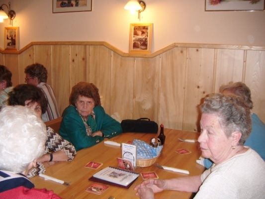 Eine Gruppe älterer Frauen sitzt um einen Holztisch in einem Restaurant. Der Tisch hat Karten, eine Flasche und einen Korb. Die Wand hinter ihnen ist aus Holz mit Bilderrahmen.