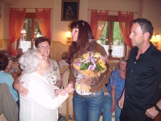 Eine Frau mit einem Blumenstrauß steht zwischen einer älteren Frau und einem Mann in einem Raum mit roten Vorhängen und einem Bilderrahmen an der Wand.