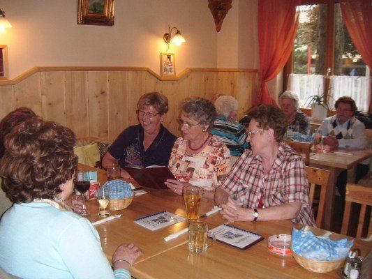 Eine Gruppe von Frauen sitzt an einem Tisch in einem Restaurant, lächelt und unterhält sich. Der Tisch hat Menüs, Gläser und Servietten. Das Restaurant hat Holzwände und ein Fenster mit Vorhängen.