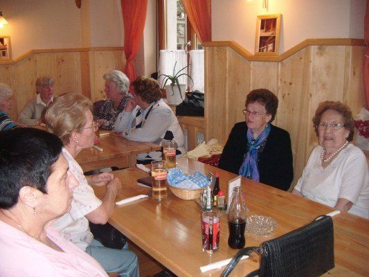 Eine Gruppe älterer Frauen sitzt in einem Restaurant um einen Tisch herum und unterhält sich. Der Tisch ist mit Gläsern, einem Korb und verschiedenen Gewürzen gedeckt. Die Wände sind aus Holz und es gibt ein Fenster mit Vorhängen.