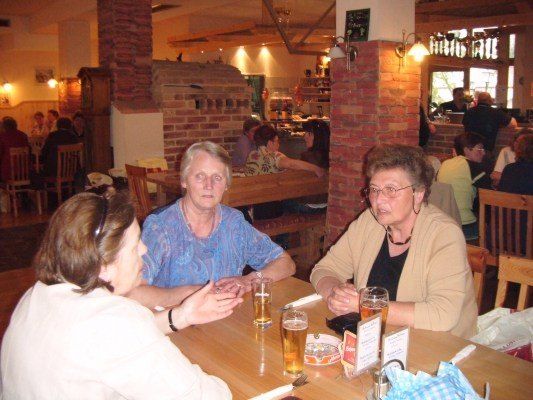 Drei Frauen sitzen an einem Tisch in einem Restaurant, zwei mit Biergläsern, in einer Unterhaltung verwickelt. Dahinter ist eine Ziegelwand und eine Theke mit einem Tafel sichtbar.