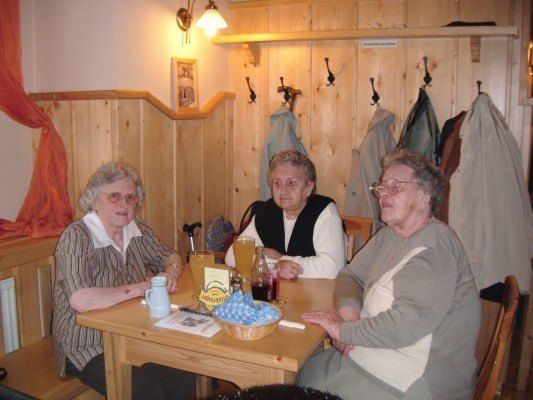 Drei ältere Frauen sitzen an einem Holztisch, lächelnd, mit Getränken und einem Korb mit Essen vor ihnen. Hinter ihnen sind hölzerne Wände mit hängenden Mänteln und einem Bilderrahmen geschmückt.