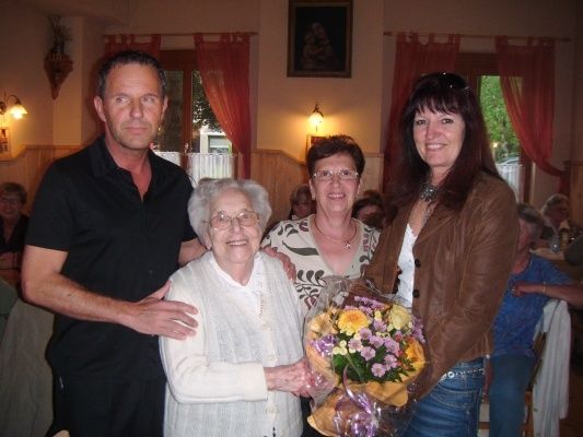 Eine Gruppe von Menschen in einem Raum, wobei eine ältere Frau einen Blumenstrauß hält und zwei Frauen neben ihr stehen. Ein Mann steht hinter der älteren Frau.