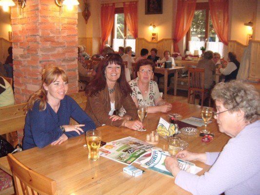 Vier Frauen sitzen an einem Tisch in einem Restaurant, lächeln und unterhalten sich. Der Tisch hat Biergläser, ein Spielbrett und andere Gegenstände. Im Hintergrund ist eine Ziegelwand und ein Fenster mit roten Vorhängen zu sehen.
