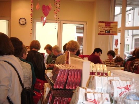 Eine Gruppe von Menschen steht in einer Schlange in einem Geschäft, möglicherweise um Geschenke zu kaufen. Der Laden hat eine Theke mit Schachteln mit Pralinen und anderen Artikeln. Es hängen herzförmige Dekorationen an der Wand.