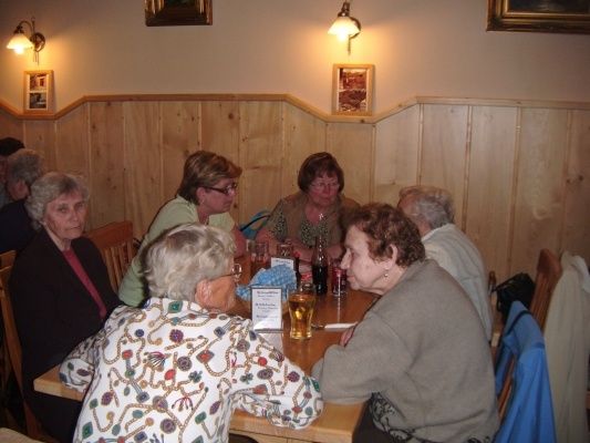 Eine Gruppe älterer Frauen sitzt in einem Restaurant um einen Tisch herum, mit Getränken und einer Speisekarte vor ihnen. Der Raum hat Holzwände und Bilderrahmen an der Wand.