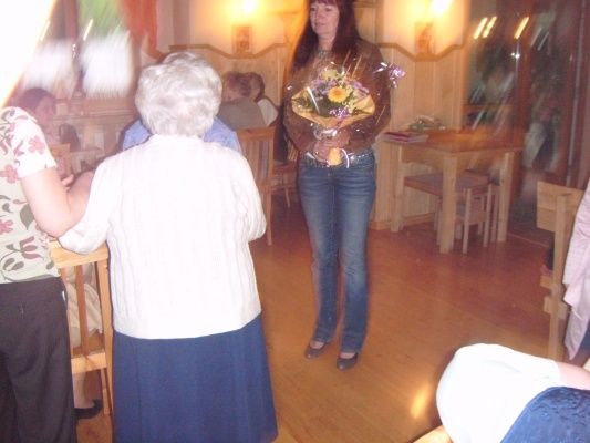 Eine Frau mit einem Blumenstrauß steht in einem Raum mit Holzböden und Möbeln, während eine andere Frau zusieht.