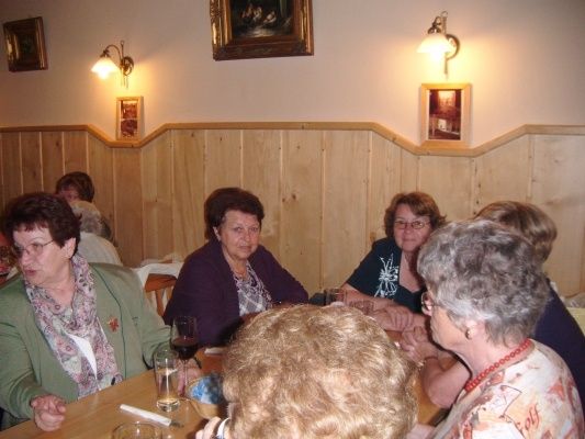 Eine Gruppe älterer Frauen sitzt in einem Restaurant um einen Tisch, einige tragen Brillen und Schals, und es hängen Bilderrahmen an der Wand.