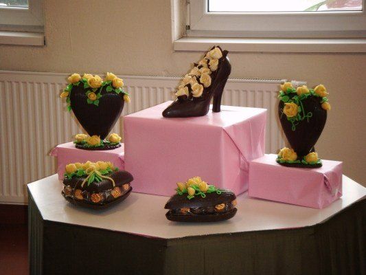 Ein Tisch zeigt fünf Schokoladenfiguren, darunter ein Schuh und vier herzförmige Stücke, jeweils mit gelben Blumen verziert. Der Schuh steht auf einem rosa Kasten, während die Herzen auf kleineren rosa Kästen stehen. Im Hintergrund befindet sich eine weiße Wand und ein Fenster.