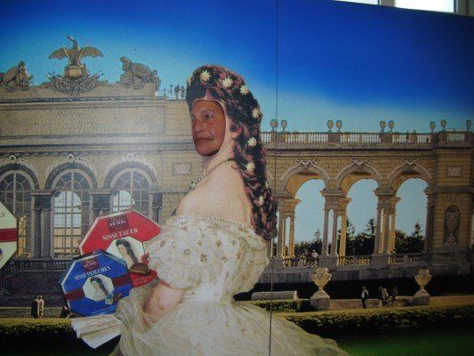 Eine Person mit einer Ausschnitt des Gesichts einer Frau, die eine Schachtel Sini Taler Schokoladen vor einem großen Palast hält.