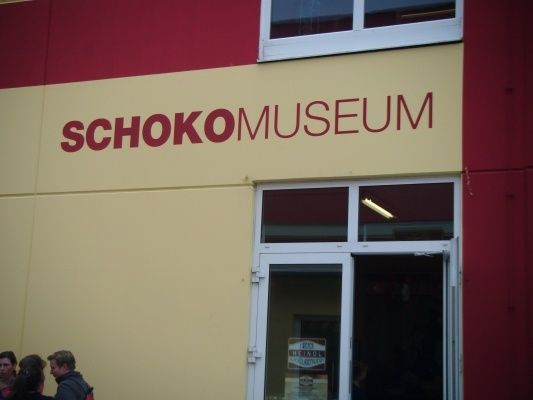 Der Eingang des Schoko Museums, mit einer rot-gelben Wand, auf der der Name des Museums in fetten roten Buchstaben steht. Eine Glastür mit weißem Rahmen ist geöffnet, und Menschen stehen davor.