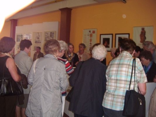 Eine Gruppe von Menschen steht in einem Raum, wahrscheinlich in einer Kunstgalerie. Sie betrachten die Gemälde an der Wand. Der Raum hat gelbe Wände und eine weiße Decke.