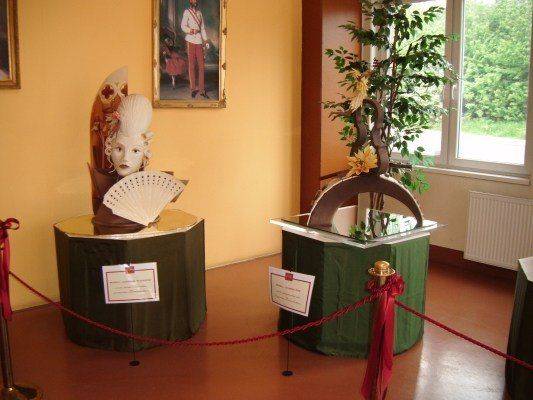 Zwei dekorative Skulpturen sind auf grünen Podesten mit roten Bändern ausgestellt. Eine Skulptur zeigt den Kopf einer Frau mit einem Fächer, während die andere eine baumartige Struktur darstellt. Beide sind von einem roten Seilzaun umgeben. Dahinter befindet sich eine Wand mit einem gerahmten Porträt und einem Fenster mit Blick auf Bäume draußen.