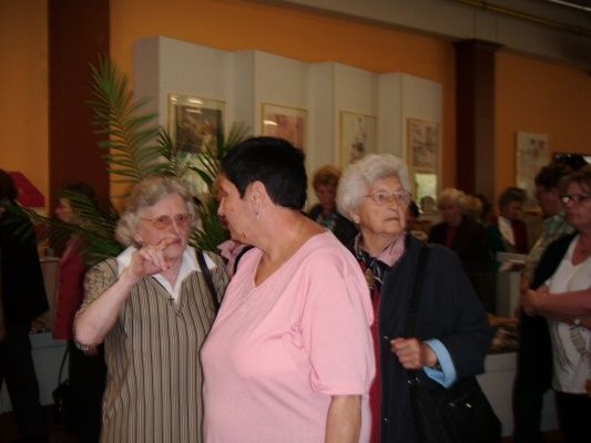 Drei ältere Frauen stehen in einem Raum, wahrscheinlich bei einer Veranstaltung, wobei eine Frau eine Brille und ein gestreiftes Kleid trägt, eine andere ein rosa Shirt und die dritte eine blaue Jacke. Hinter ihnen versammeln sich mehrere Menschen, und der Raum ist mit gerahmten Bildern an der Wand dekoriert.