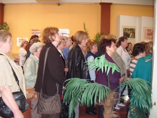 Eine Gruppe von Frauen, größtenteils älterer, steht in einem Raum, wahrscheinlich bei einer Veranstaltung. Sie schauen in die gleiche Richtung, möglicherweise auf eine Ausstellung oder Präsentation. Der Raum hat gelbe Wände, Topfpflanzen und gerahmte Bilder an der Wand.