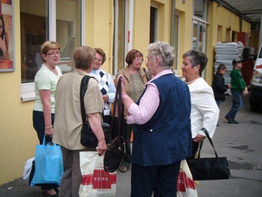 Eine Gruppe von Frauen mit Einkaufstaschen steht vor einem gelben Gebäude, eine Frau hält eine schwarze Tasche, während eine andere eine blaue Tasche hält. Ein Kind geht im Hintergrund vorbei.