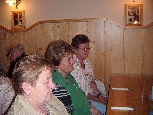 Vier ältere Frauen sitzen in einer hölzernen Kabine und schauen nach vorne. Die Frau auf der linken Seite trägt ein beiges Oberteil. Die Frau neben ihr trägt eine grüne Jacke. Die beiden Frauen rechts tragen weiße und gestreifte Oberteile.