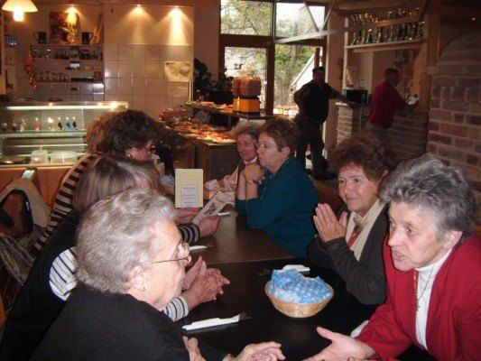 Eine Gruppe älterer Frauen sitzt in einem Restaurant um einen Tisch herum, klatscht und lächelt. Ein Korb mit blauen Bonbons steht auf dem Tisch.