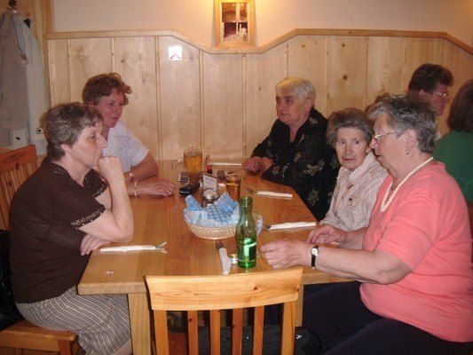 Eine Gruppe älterer Frauen sitzt in einem Restaurant um einen Holztisch herum und führt ein Gespräch. Der Tisch ist mit Gläsern, einem Korb und einer grünen Flasche gedeckt. Holzstühle umgeben den Tisch, und eine Holzwand mit einem Bilderrahmen ist im Hintergrund.