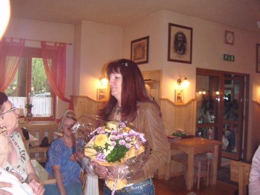 Eine Frau hält einen Blumenstrauß in einem Restaurant, wo andere Leute an Tischen sitzen. Die Wände sind mit gerahmten Bildern und einer Uhr dekoriert.