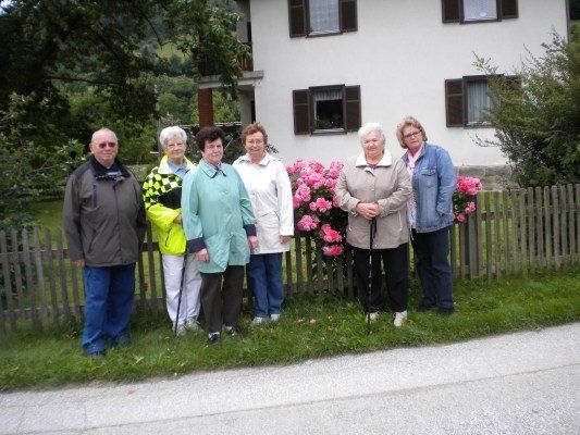 Eine Gruppe älterer Menschen steht vor einem Haus mit rosa Blumen und einem Holzzaun.