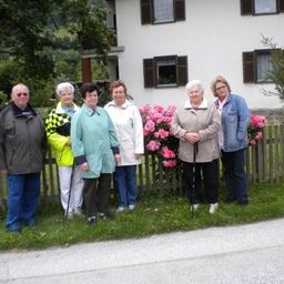 Eine Gruppe älterer Menschen steht vor einem Haus mit rosa Blumen und einem Holzzaun.