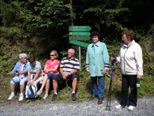 Eine Gruppe älterer Menschen sitzt auf einer Bank im Wald, einer hält eine Flasche und einen Trekkingstock. Ein Wegweiser mit Wegbeschreibungen steht hinter ihnen.